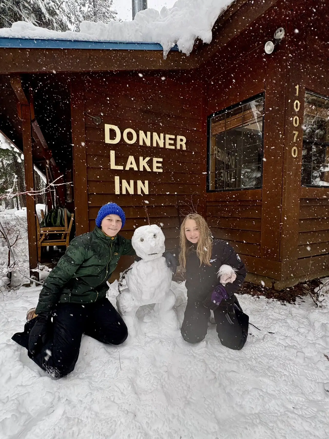 Two kids kneeling beside a snowman in front of the Donner Lake Inn sign as snow falls