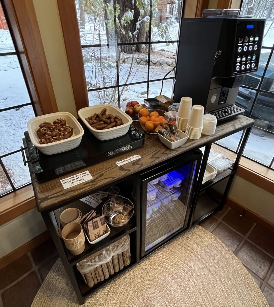 Grab-and-go breakfast setup at Donner Lake Inn with muffins, chicken sausage, fresh fruit, yogurt, and self-serve coffee by a window overlooking the pines